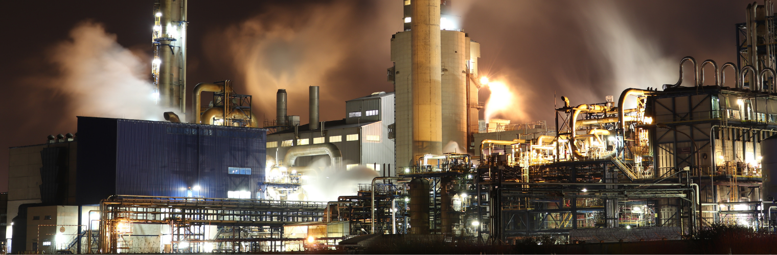 A large industrial plant illuminated at night, with smoke billowing from its towering chimneys against a dark sky.