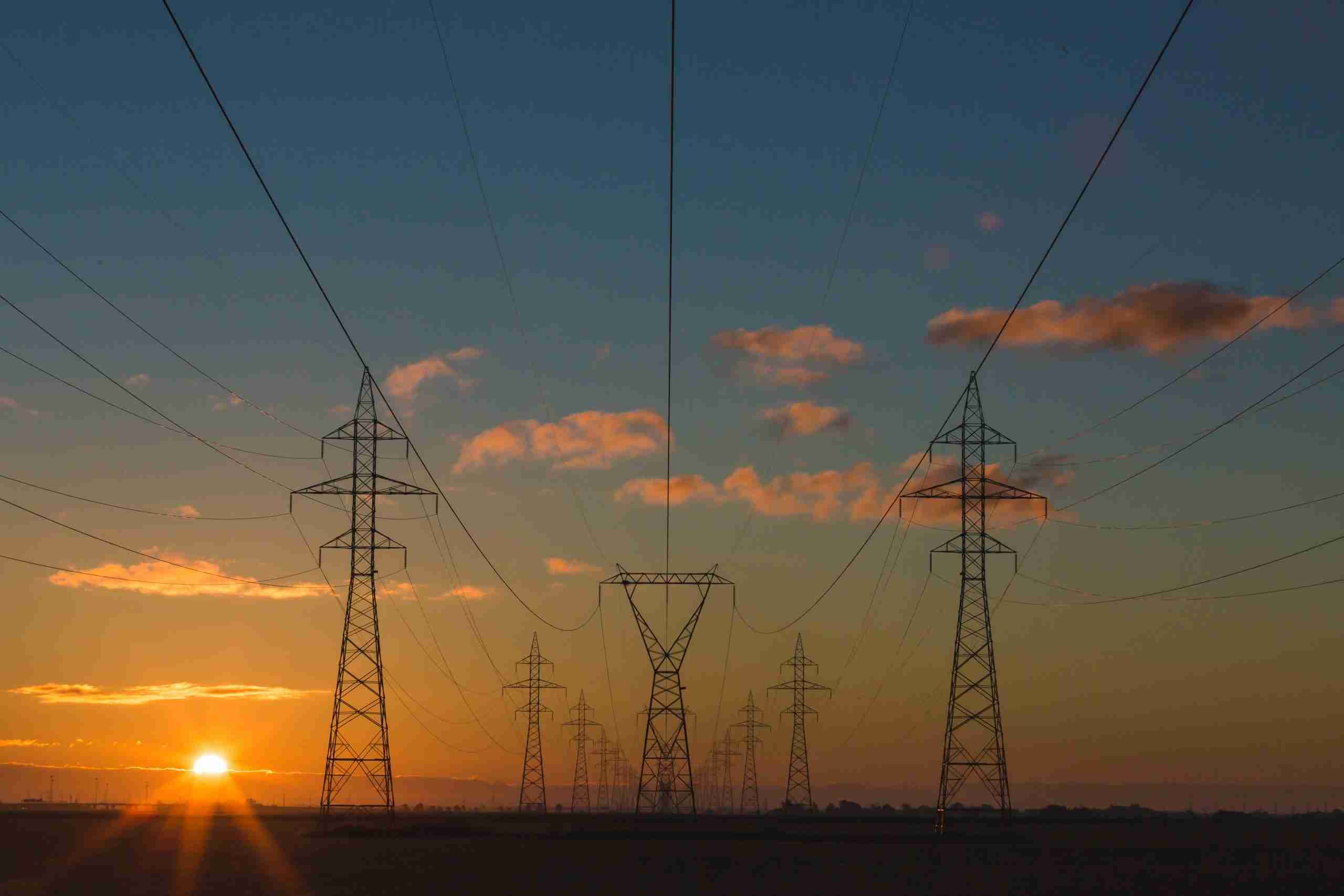A sunset casts a warm glow behind towering high voltage power lines, silhouetting their structure against the colorful sky.